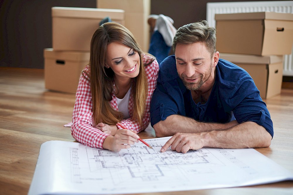 Žena s mužem leží na zemi a dívají se do papírového plánu domu, v pozadí jsou papírové krabice